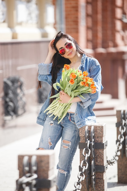 Mujer atractiva joven que sostiene las flores al aire libre. Señora urbana con ropa casual sobre fondo urbano. Hermosa mujer en la ciudad.