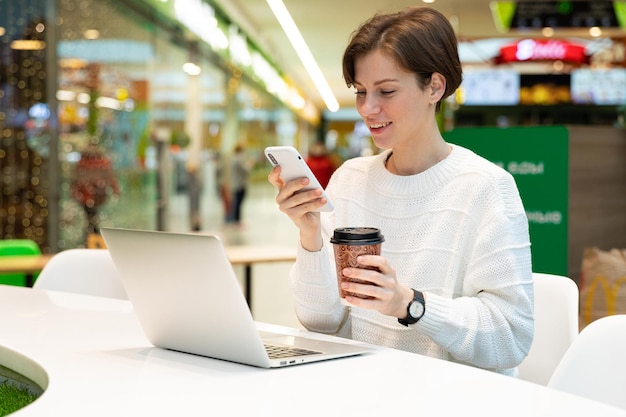 Mujer atractiva joven sentada en un centro comercial en una mesa y trabajando en una computadora portátil