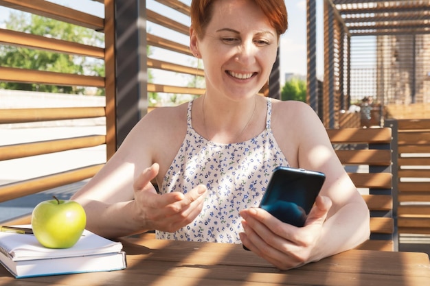 Mujer atractiva joven se sienta en la terraza y sonríe mientras mira el teléfono