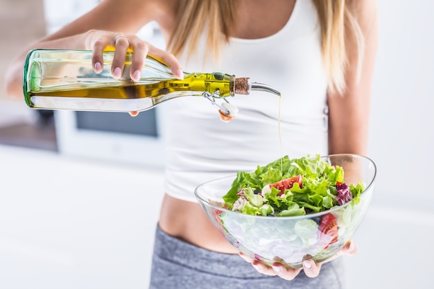 Mujer atractiva joven vertiendo aceite de oliva en la ensalada de verduras frescas.