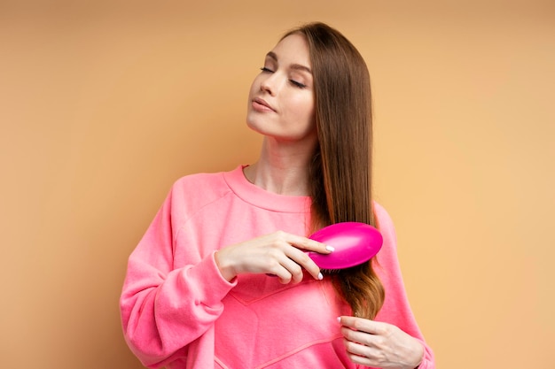 Mujer atractiva y linda con los ojos cerrados usando una sudadera rosa cepillándose el cabello con un cepillo