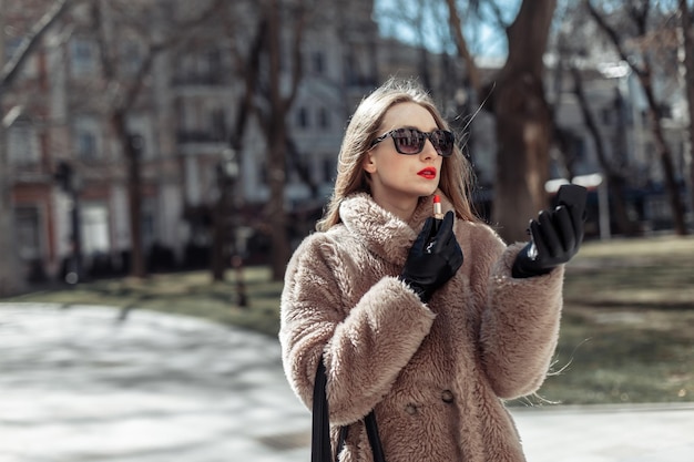 Mujer atractiva de lujo con un abrigo de piel y gafas de sol pinta sus labios con lápiz labial rojo en la ciudad