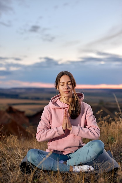 Mujer atractiva mantenga la calma, meditando sobre la estera de fitness sentado al aire libre en la naturaleza