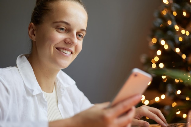 Mujer atractiva con peinado de moño en camisa blanca sentada en la mesa cerca del árbol de navidad sosteniendo un teléfono inteligente en las manos expresando emociones positivas leyendo mensajes de felicitación con año nuevo