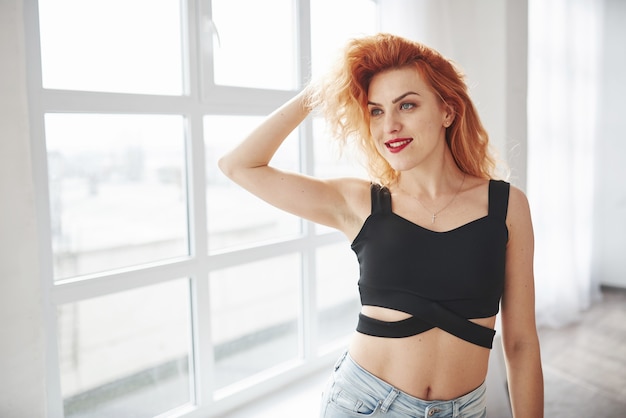 Mujer atractiva pelirroja posando en la habitación espaciosa cerca de la ventana.