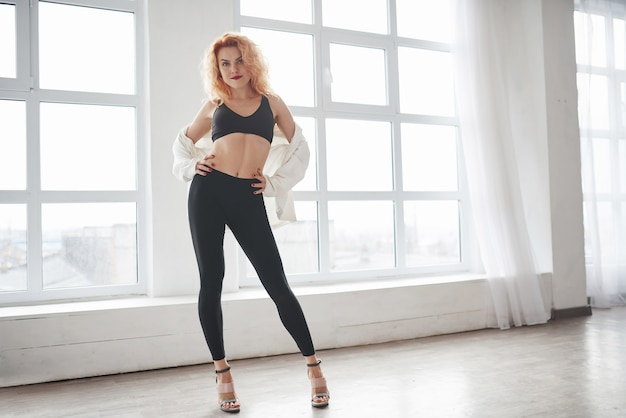 Mujer atractiva pelirroja posando en la habitación espaciosa cerca de la ventana.