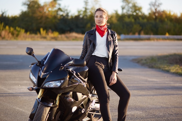 La mujer atractiva pensativa tiene una mirada pensativa en la distancia, vestida con una chaqueta de cuero, pañuelo rojo, descansa al aire libre después de conducir una motocicleta, recorre largas distancias. Recreación y concepto de personas