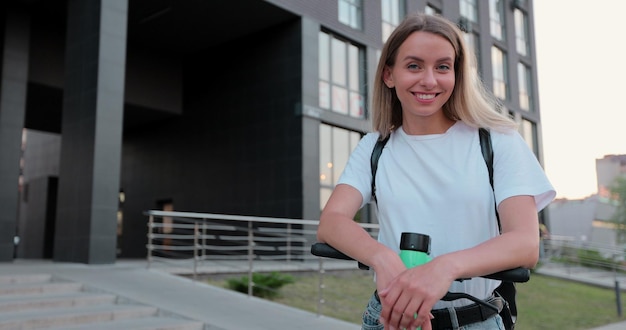 Mujer atractiva de pie con scooter eléctrico y posando para la cámara en la puesta de sol de la ciudad