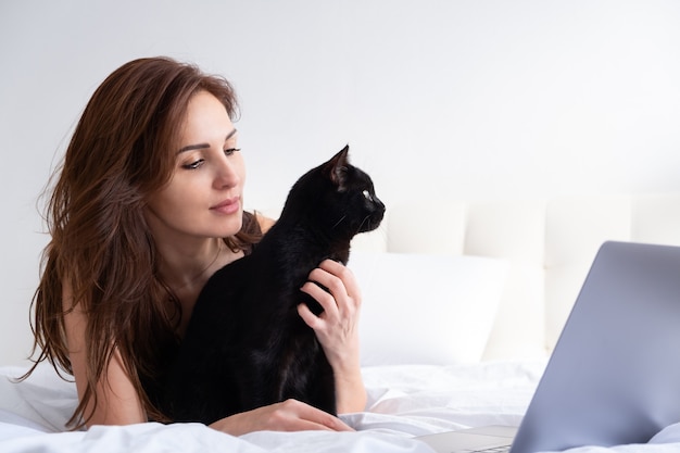 Foto mujer atractiva con portátil con su gato negro, sentando juntos en la habitación blanca. vida con mascota.