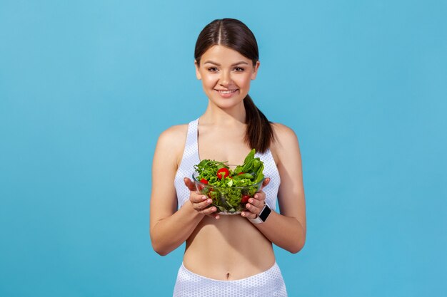 Mujer atractiva positiva sosteniendo el tazón con ensalada de verduras frescas