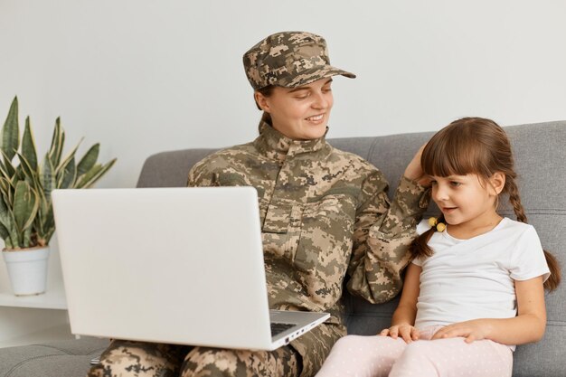 Mujer atractiva positiva con uniforme de camuflaje y gorra sentada tosiendo con su hija soldado mirando a su encantadora niña y sosteniendo una computadora portátil