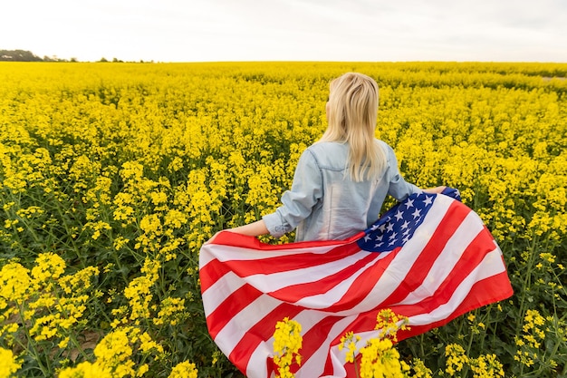 mujer atractiva que sostiene una bandera americana en el viento en un campo de colza. Paisaje de verano contra el cielo azul. Orientación horizontal.