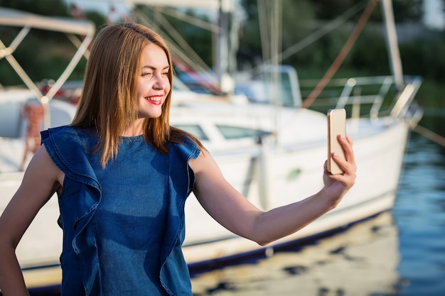 Mujer atractiva que toma la foto del selfie en el teléfono inteligente para su blog mientras está de pie en el puerto deportivo del río. Concepto de turismo y viajes.