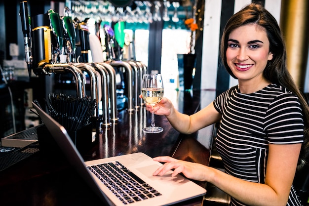 Foto mujer atractiva que usa la computadora portátil y tomando una copa de vino en un bar