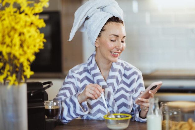 Una mujer atractiva que usa un teléfono inteligente mientras disfruta del desayuno de cereales y tiene un ritual de placer matutino en casa.