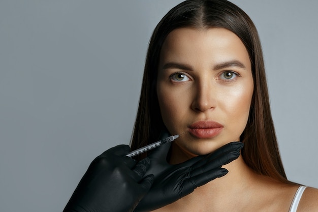 Mujer atractiva recibiendo inyección de ácido hialurónico en los labios. Foto de estudio sobre un fondo gris