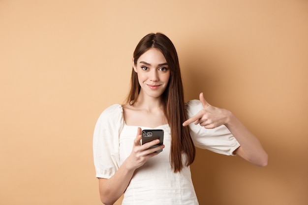 Mujer atractiva sonriendo y apuntando al teléfono inteligente, mostrando la oferta en línea en el teléfono, de pie sobre fondo beige.