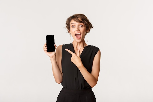 Mujer atractiva sorprendida mirando emocionado, señalar con el dedo en la pantalla del teléfono inteligente