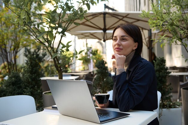 Mujer atractiva sosteniendo una taza de café y trabajando en la computadora portátil sentado en una mesa en la cafetería al aire libre en un hermoso día cálido. Negocio, concepto de trabajo a distancia