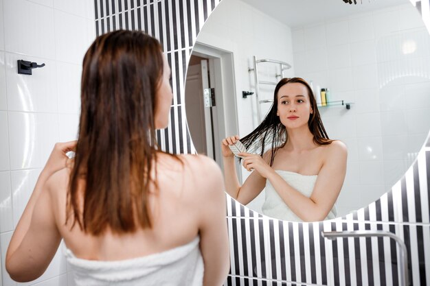 Mujer atractiva en toalla blanca con peine cepillando su cabello mojado después de ducharse en casa frente al espejo del baño Se preocupa por el cabello sano y limpio Concepto de belleza