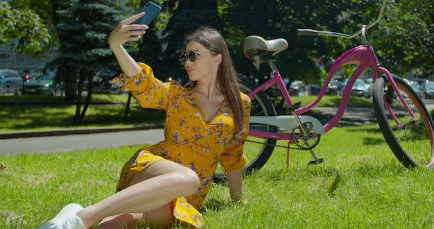 Mujer atractiva tomando un selfie en un teléfono inteligente sentado en el parque en el césped. Close-up Hermosa mujer sentada en el césped en el parque cerca de la bicicleta y tomando un selfie en el teléfono inteligente.