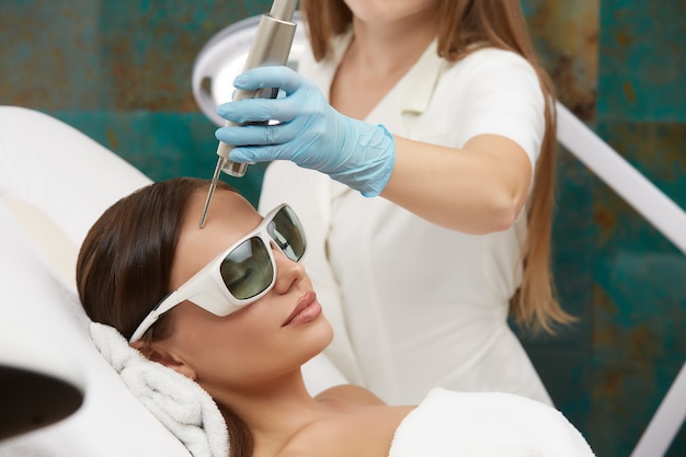 Mujer atractiva con tratamiento facial con láser en salón de belleza por cosmetóloga profesional
