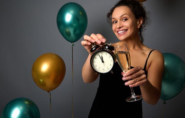 Mujer atractiva en vestido negro de noche sostiene una flauta de vino espumoso y muestra un despertador aislado sobre fondo gris con globos de aire inflados. Concepto de Navidad y año nuevo, aniversario.