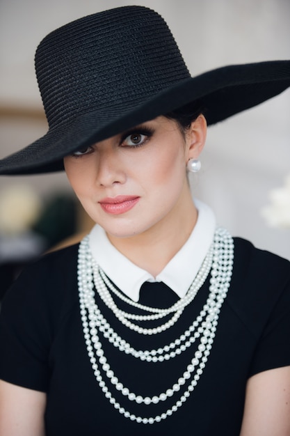 Foto mujer atractiva con vestido negro, sombrero y perlas, sentado en la silla