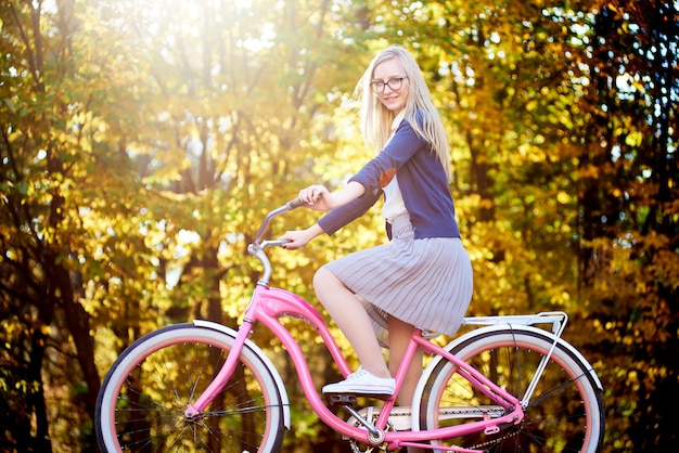 Mujer atractiva del viajero que monta en la bicicleta moderna de la señora rosada encendida por el parque del sol del otoño en fondo colorido brillante de los árboles del bokeh de oro.