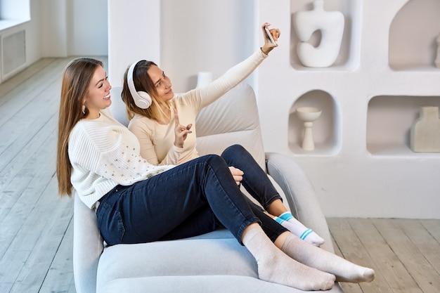 Mujer en auriculares inalámbricos hace selfie con hermana por teléfono en interiores