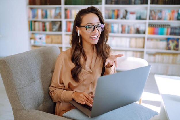 Mujer con auriculares inalámbricos llamando en una computadora portátil hablando por cámara web sentada en un café moderno Educación