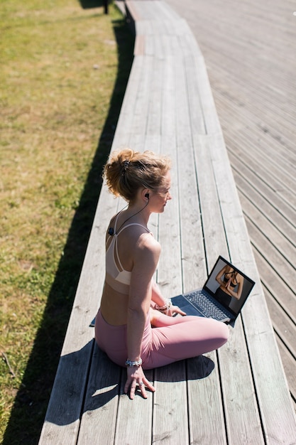 Mujer en auriculares sentado en pose de yoga al aire libre en el paseo marítimo de madera viendo videos en la computadora portátil