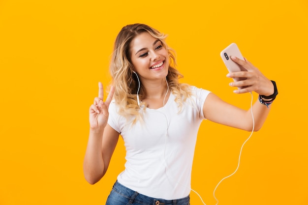 Mujer con auriculares sonriendo y tomando fotos selfie en teléfono celular, aislado sobre pared amarilla