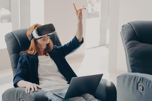 Mujer en auriculares VR apuntando con el dedo tocando objetos 3d mientras trabaja de forma remota en una computadora portátil
