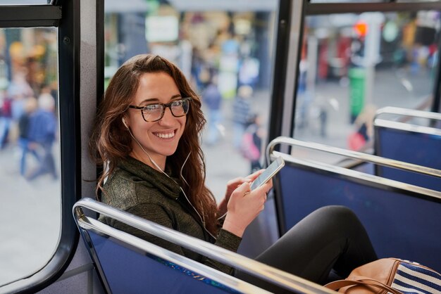 Mujer autobús y auriculares con teléfono para transporte de retratos y sonrisa con redes sociales Person música y teléfono inteligente para escuchar con suscripción de transmisión de audio en aplicación móvil en Nueva York