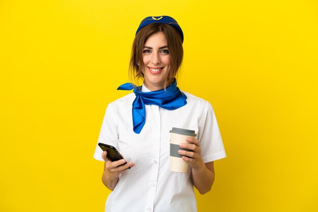 Mujer azafata de avión aislada sobre fondo amarillo sosteniendo café para llevar y un móvil