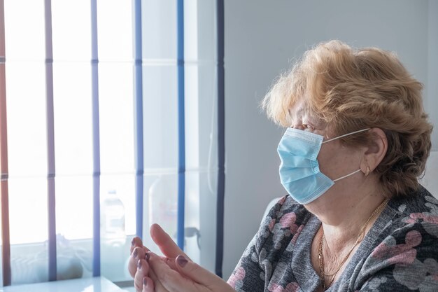 Mujer en azul máscara protectora médica quedarse en casa y mirar por la ventana. Cuarentena