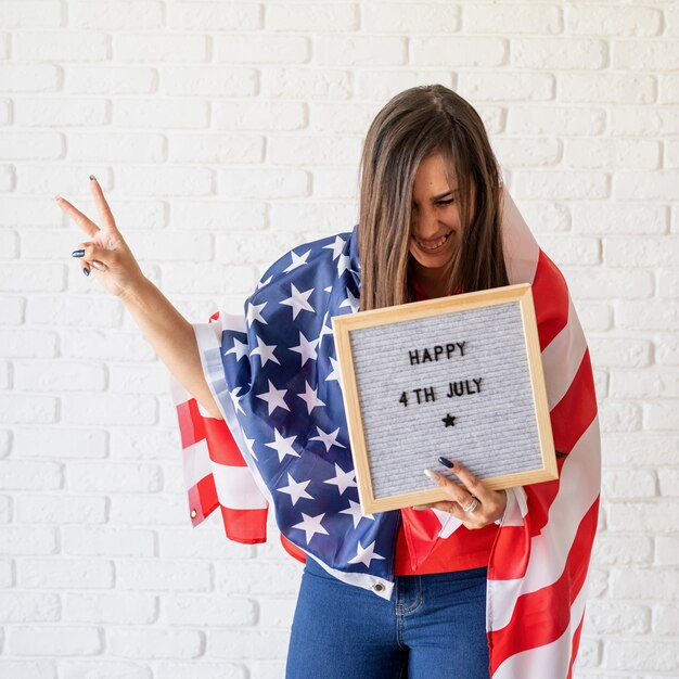 Mujer con bandera americana sosteniendo el tablero de letras con palabras feliz 4 de julio