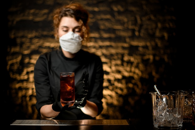 Mujer barman en máscara médica y guantes negros presentan vidrio con cóctel