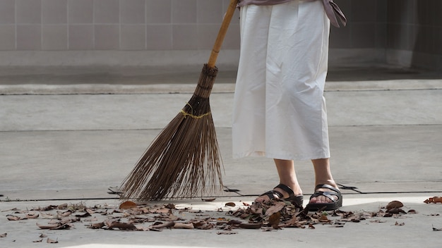 Mujer barriendo las hojas secas en el piso de cemento con una escoba de madera larga y manteniendo limpio al aire libre