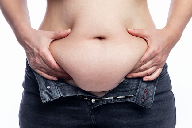 Una mujer con una barriga gorda en jeans con una bragueta desabrochada toca la grasa Cuerpo con sobrepeso agradable Primer plano Aislado sobre fondo blanco Vista frontal