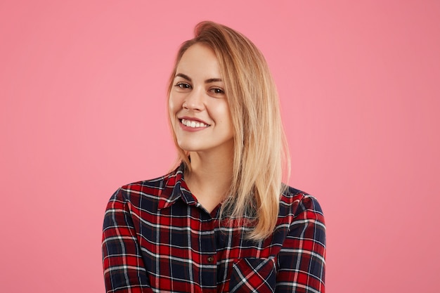 Mujer bastante adorable con una sonrisa agradable mira directamente a la cámara, vestida con una camisa a cuadros, aislada sobre la pared rosa. Sonriente joven amigable se regocija por su éxito, posa en interiores