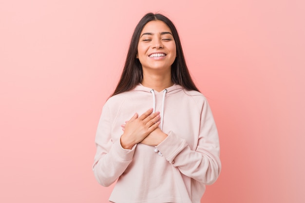 La mujer bastante árabe joven que lleva una mirada casual del deporte que ríe manteniendo las manos en el corazón, concepto de felicidad.