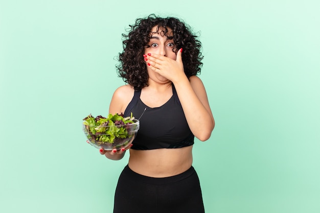 Mujer bastante árabe que cubre la boca con las manos con un sorprendido y sosteniendo una ensalada. concepto de dieta