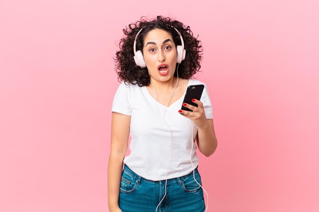 Mujer bastante árabe que parece muy conmocionada o sorprendida con auriculares y un teléfono inteligente