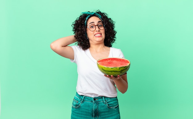 Mujer bastante árabe que se siente estresada, ansiosa o asustada, con las manos en la cabeza y sosteniendo una sandía. concepto de verano