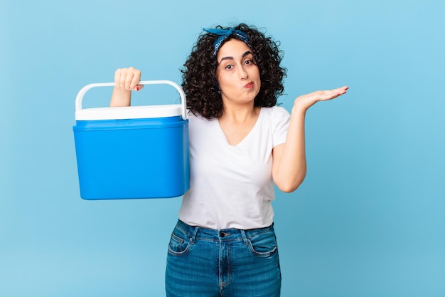 Mujer bastante árabe que se siente perpleja y confundida y dudando y sosteniendo un refrigerador portátil