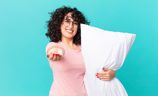 Mujer bastante árabe sonriendo felizmente con amigable y ofreciendo y mostrando un concepto. en pijama y sosteniendo una almohada