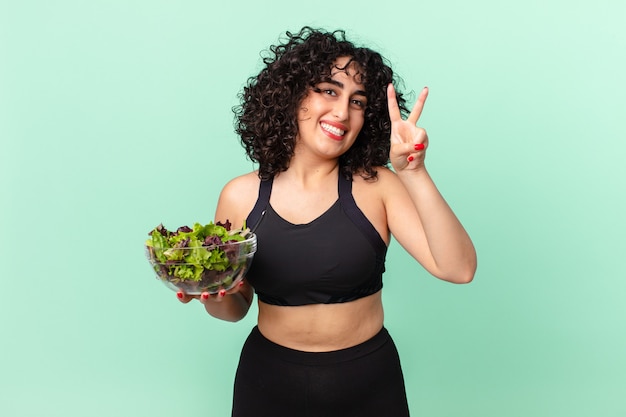 Mujer bastante árabe sonriendo y mirando amigable, mostrando el número dos y sosteniendo una ensalada. concepto de dieta