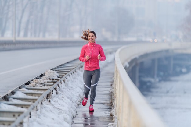 Mujer bastante caucásica con cola de caballo vestida con ropa deportiva corriendo en el puente en invierno. Concepto de estilo de vida saludable